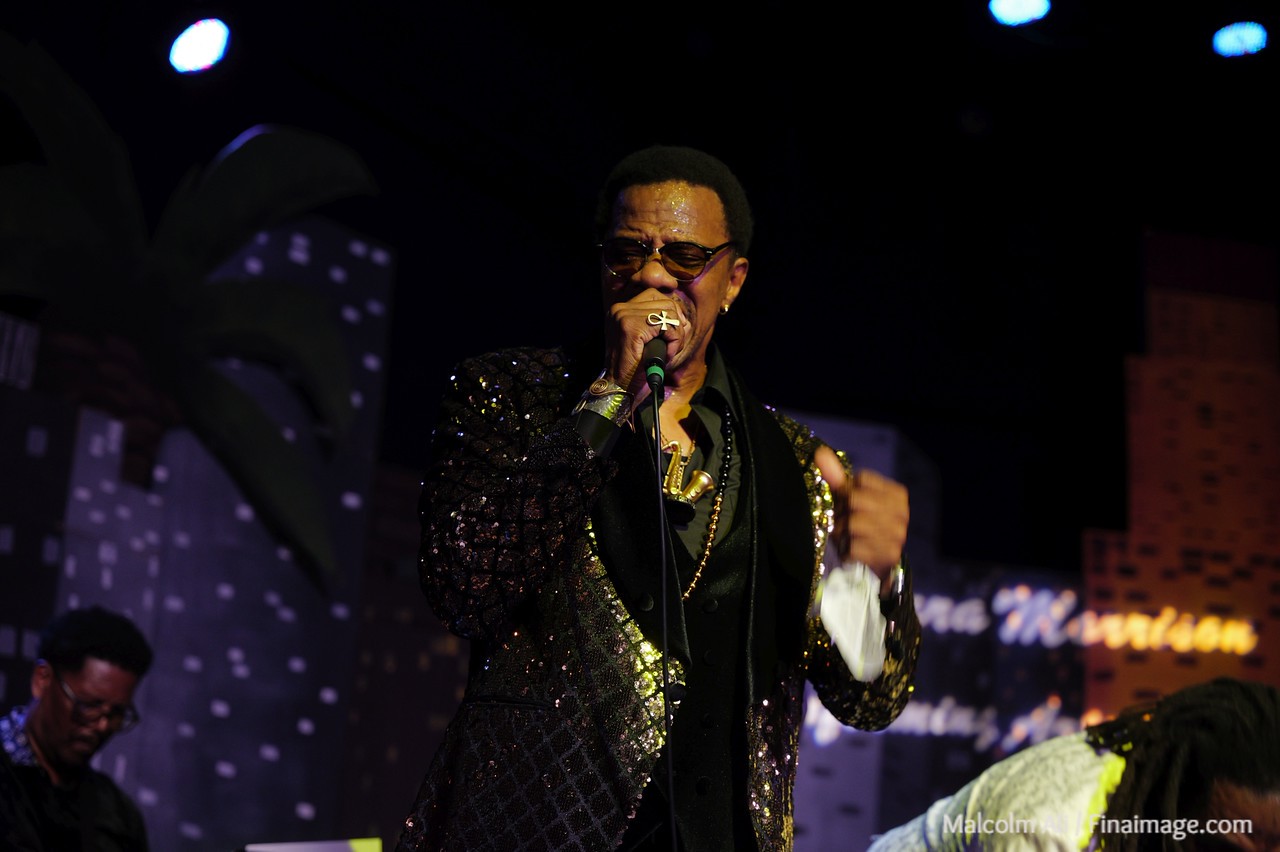 Performer singing into microphone on stage, wearing sunglasses and a sparkly jacket, with a dark background and stage lights.